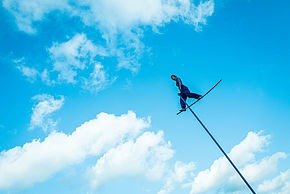 À plusieurs mètres de hauteur, une femme, habillée tout en bleu, se tient debout sur une tige bleue. En arrière fond, le ciel et les nuages.