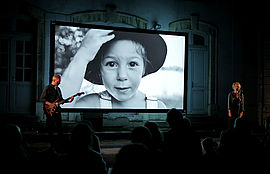 Sur une scène, un guitariste et une comédienne devant un micro. Derrière eux, un écran avec une petite fille qui porte un chapeau et une salopette. Devant eux le public. - Agrandir l'image (fenêtre modale)