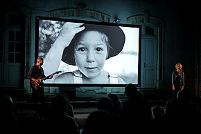 "Cavale", compagnie Les Mangeurs d’étoiles, 6ème édition de la Biennale Urbaine de Spectacles, 2022, Pantin, Sur une scène, un guitariste et une comédienne devant un micro. Derrière eux, un écran avec une petite fille qui porte un chapeau et une salopette. Devant eux le public.